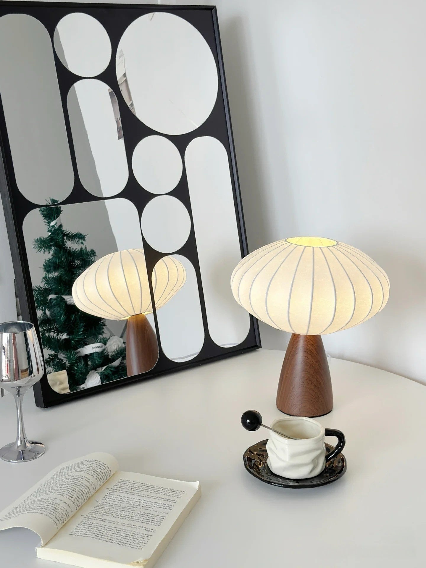 Mid-century modern table lamp with pleated fabric shade and walnut base, placed on a white desk with coffee cup, book, and decorative mirror