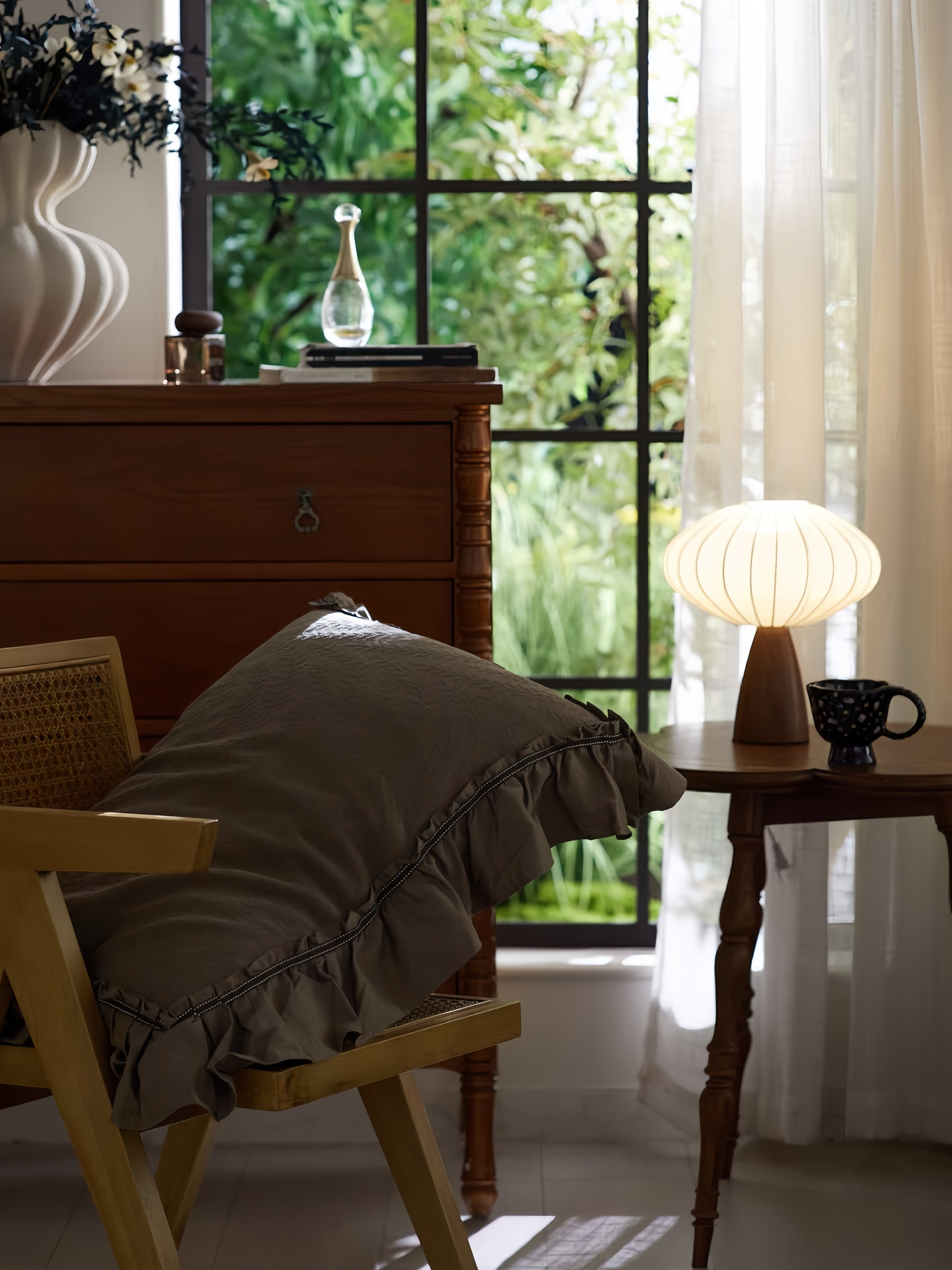 Cozy mid-century modern corner with cane chair, wooden side table, pleated fabric table lamp, and greenery view through large window
